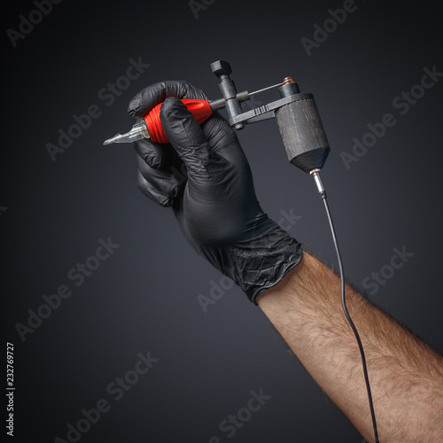 Tattooist hands in black gloves with tattoo machine on dark gray background. photo