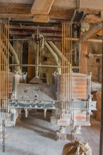Portrait format of traditional flour mill equipment, covered in flour from milling, with shutes feeding mill with organic grain and a full bag of flour on floor in front. © Marion Smith (Byers)