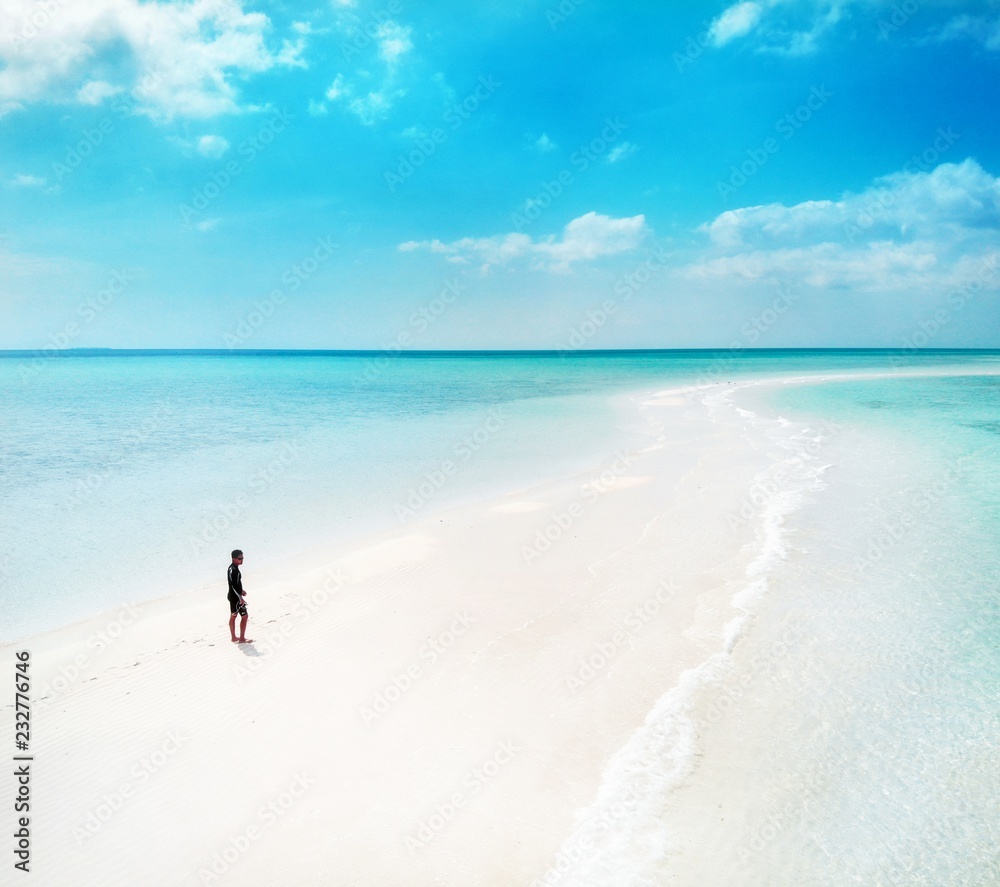 Panoramic aerial photo of a young man walking at the beach by the seashore in small island with white sand and crystal clear blue turquoise water