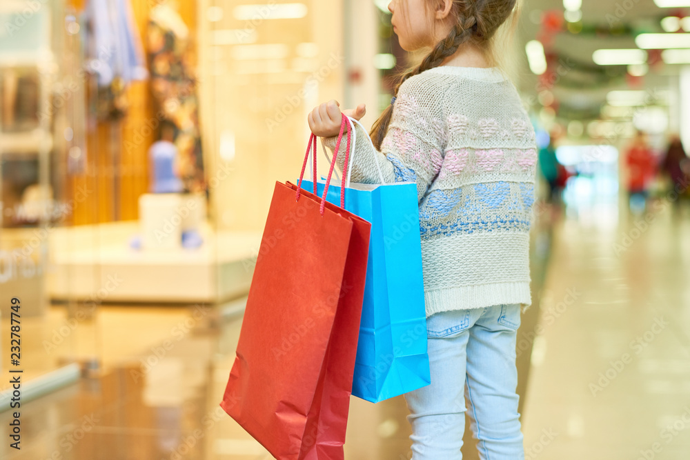 Rear view portrait of cute little girl holding big paper shopping bag standing against window display, copy space Rear view portrait of cute little girl holding big paper shopping bag standing against window display, copy space