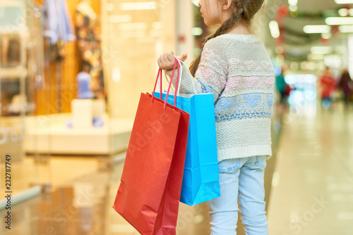 Rear view portrait of cute little girl holding big paper shopping bag standing against window display, copy space