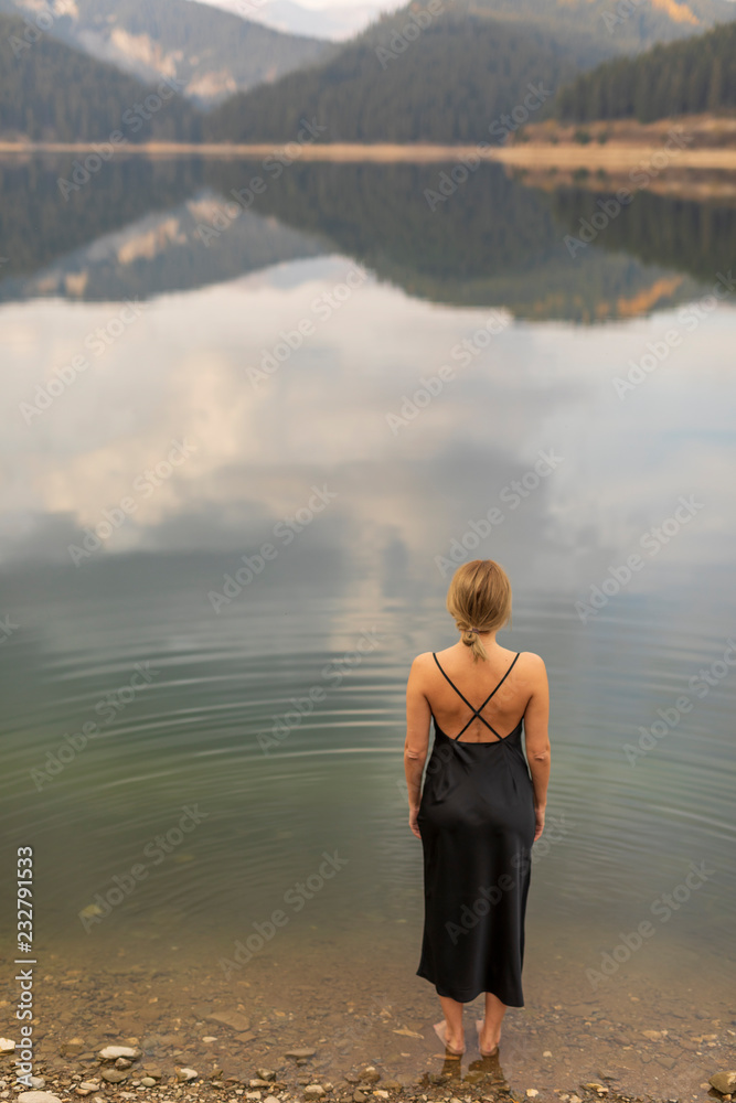 Attractive blond fashion model from behind in a front of a lake. Rear view of elegant woman in black evening dress. Attractive blond fashion model from behind in a front of a lake. Rear view of elegant woman in black evening dress.