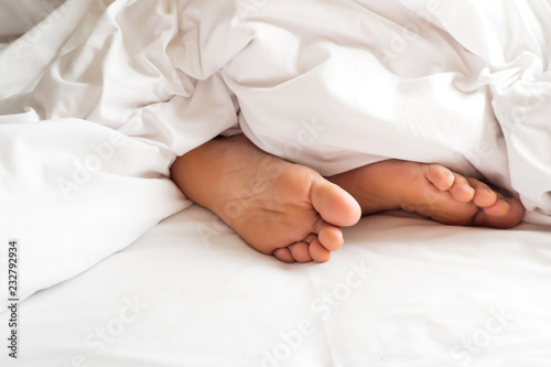 Sole of woman foot in messy blanket on bed. White pillow with blanket on bed unmade. Concept of relaxing after morning.