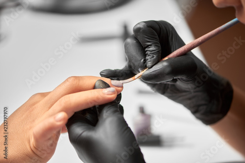 Process of making manicure in beauty salon. Close-up of professional beautician hands in black gloves applying pastel varnish on client woman nails using brush on light blurred copy space background.