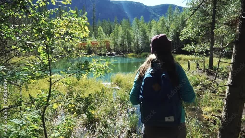 Young woman tourist is coming near on a picturesque blue lake in the mountains in sunny day. Back view Young woman tourist is coming near on a picturesque blue lake in the mountains in sunny day. Back view