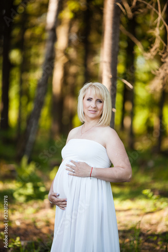 Portrait of beautiful pregnant woman in sheer long white maternity dress looking dreamy in the middle of forest © Cimermane