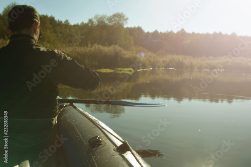 A man in black clothes and a cap swims in an inflatable boat with oars on the river on a clear autumn day in the rays of the sun.