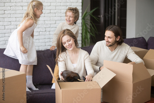 Young first time buyers, happy family in living room. Parents sitting on couch and unpacking cardboard boxes, little kids play jump on sofa. Buy real estate, relocating at new house, mortgage concept