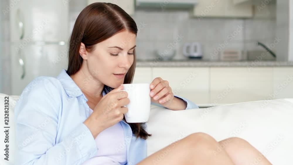 Young brunette woman with brown eyes relaxing sitting on couch at home in the morning. Pretty female housewife drinking tasty coffee or tea from white cup in living room. Kitchen on background Young brunette woman with brown eyes relaxing sitting on couch at home in the morning. Pretty female housewife drinking tasty coffee or tea from white cup in living room. Kitchen on background