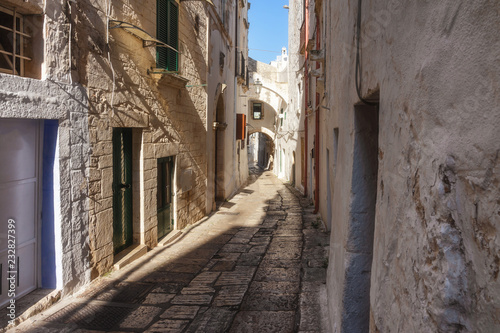 A narrow street surrounded by vintage  stone architecture and with an arch at the end in the old town area of the town of Ostuni in the Puglia region of Italy on a summer day with a clear blue sky