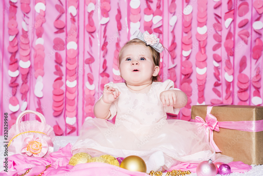 Fototapeta premium Cute little baby girl sits on a soft blanket in a pink dress with a Christmas balls, party gift and handbag, in a festive pink interior. New Year and Christmas holidays.