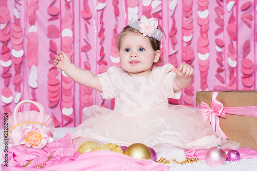 Cute little baby girl sits on a soft blanket in a pink dress with a Christmas balls, party gift and handbag, in a festive pink interior. New Year and Christmas holidays.
