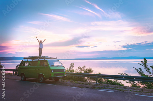 Young beautiful girl standing on the roof of old timer classic camper van parked by the sea with amazingly colorful sunset sky, hands wide open free photo