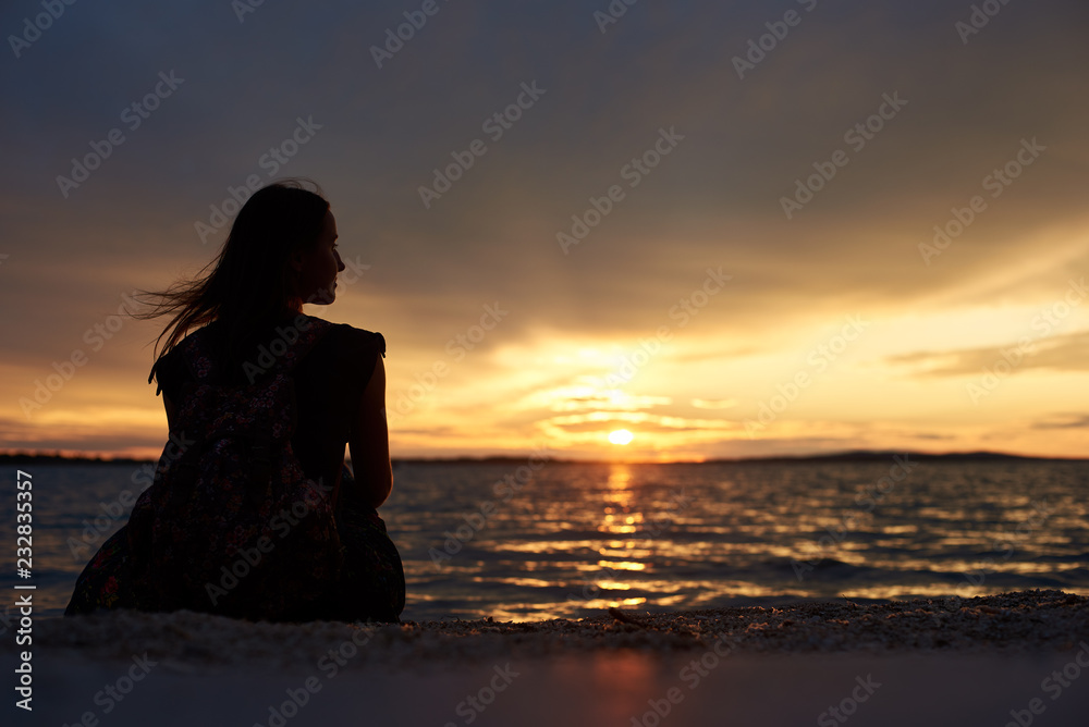 Back view silhouette of tourist woman with backpack sitting alone on seashore at water edge, enjoying beautiful view of sunset on dark evening sky background. Tourism and vacations concept.