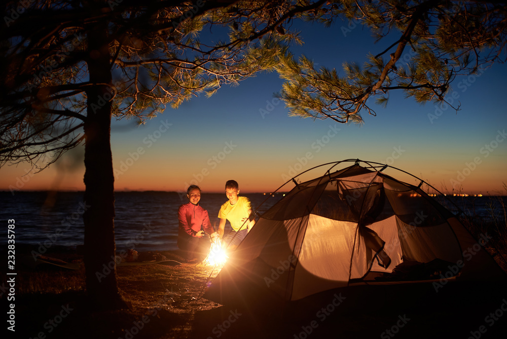 Night camping at sea. Tourist tent under tree and young couple, man and  woman at campfire against evening sky and orange glow on horizon. Tourism,  happy relations and active lifestyle concept. Photos