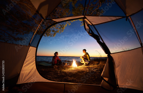 View from inside tourist tent at sunset. Young tourist romantic family, man and woman sitting at bonfire making supper on lake shore on blue evening sky and crystal blue clear lake water background.