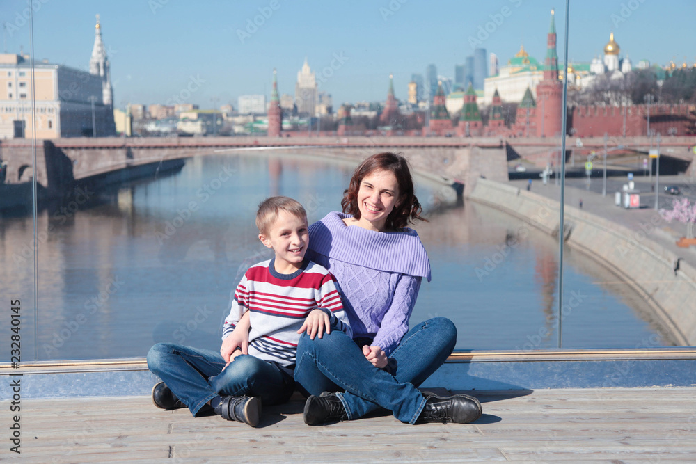 Mom and young son sitting on glass bridge on a background of the river. Traveling with kids concept