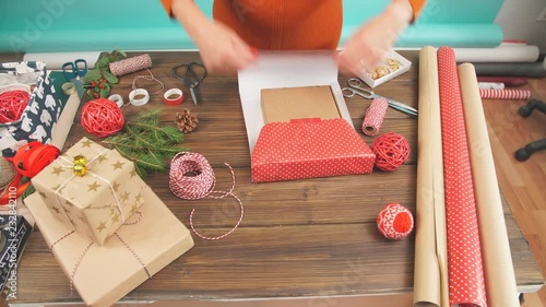 Female hands wrapping Christmas gift in craft ecological paper on dark wooden backround with decorating elements, colourfil twine stripes and pack paper rolls, Christmas or New year DIY packing. photo