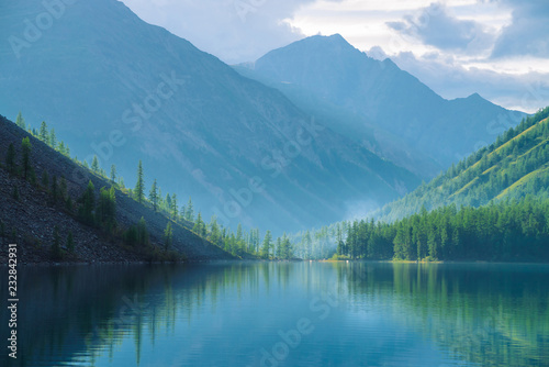 Ghostly mountain lake in highlands at early morning. Beautiful misty mountains reflected in calm clear water surface. Smoke of campfires. Amazing atmospheric foggy landscape of majestic nature.