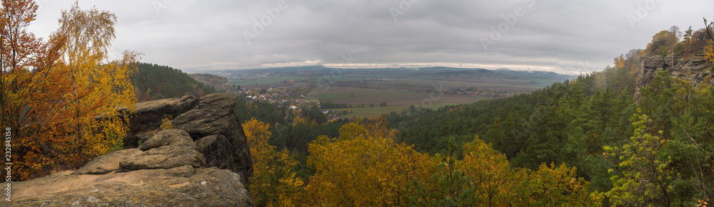 Panoramic view from sandstone pillars view-point at nature reserve Cesky Raj with autumn colored deciduous and coniferous tree forest and green hills, moody blue sky