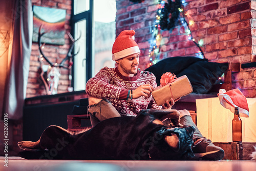 A stylish man holding a gift box while sitting with his cute dog in a decorated living room at Christmas time.
