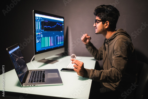 Young indian man pointed on coin looking on display with bitcoin cryptocurrency market in dark office