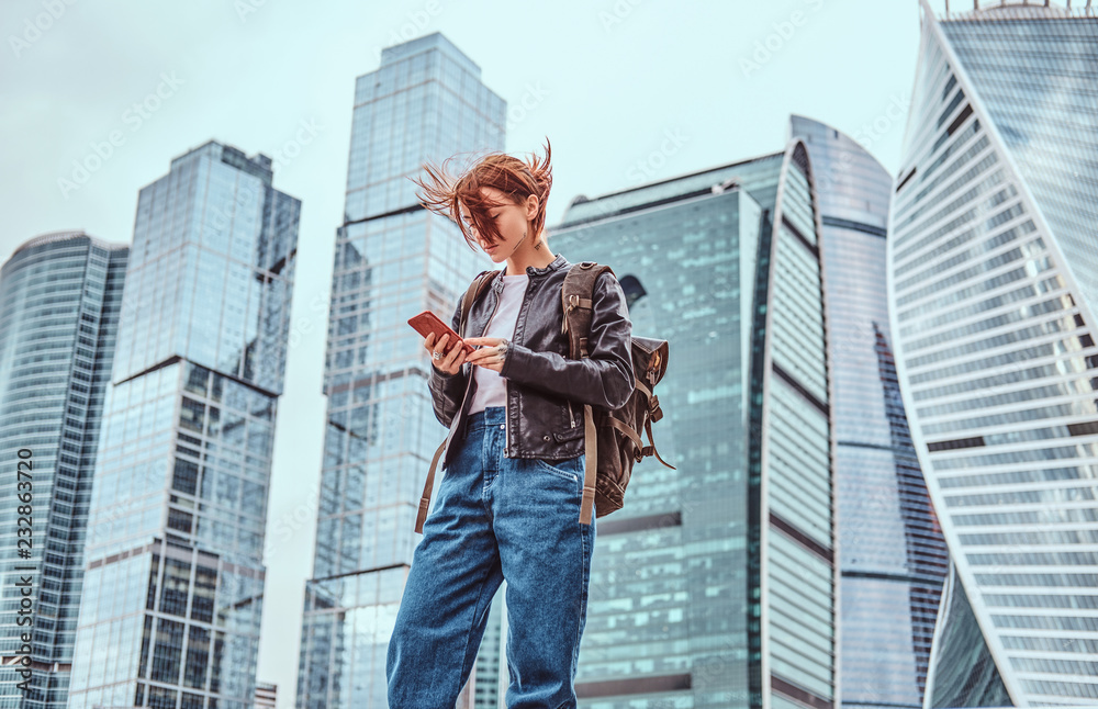 Trendy dressed redhead student girl with tattoos on her face using a smartphone in front of skyscrapers in Moskow city.