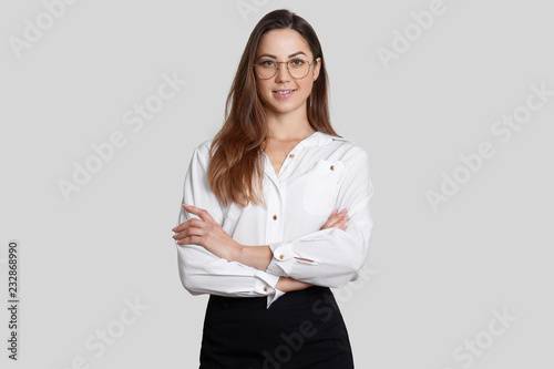 Cheerful self confident businesswoman wears white blouse and black skirt, keeps arms folded, wears round spectacles, poses against white background, listens interlocutor, dressed in formal clothes