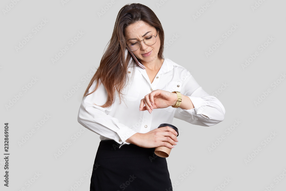 Busy young female has telephone conversation, hurries at work, looks at watch, holds aromatic takeaway coffee, dressed in formal clothes, isolated over white background. Business and work concept Busy young female has telephone conversation, hurries at work, looks at watch, holds aromatic takeaway coffee, dressed in formal clothes, isolated over white background. Business and work concept