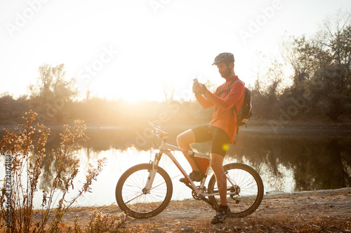 Smiling young man sitting on a mountain bike by the lake, resting and using a smart phone. Healthy and active lifestyle concept