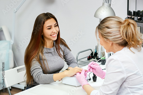 A nail artist does a manicure to a client. Brunette customer happy and smiling.