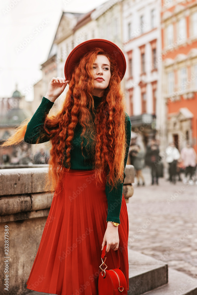 Street fashion portrait of young beautiful redhead woman with freckles,  very long curly hair, wearing green turtleneck, wrist watch, orange hat,  pleated skirt, holding stylish red round suede handbag Stock Photo |