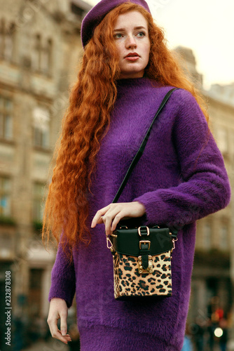 Young beautiful fashionable redhead woman with freckles, long curly hair,  wearing purple sweater, beret, holding stylish bag with animal, leopard  print. Model posing in street of european city Stock Photo | Adobe