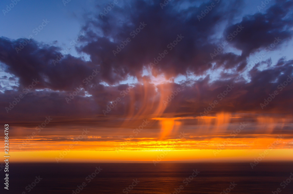 Brilliant orange sunset highlights streaks of rain falling from purple clouds over the Pacific Ocean along California's Big Sur coast