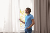 Young man standing near window with open curtains at home