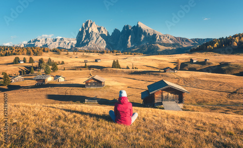 Sitting woman on the hill looking in meadows and mountains at sunset in autumn. Alpe di Siusi, Dolomites, Italy. Landscape with girl in red jacket, field with yellow grass, wooden houses and rocks 