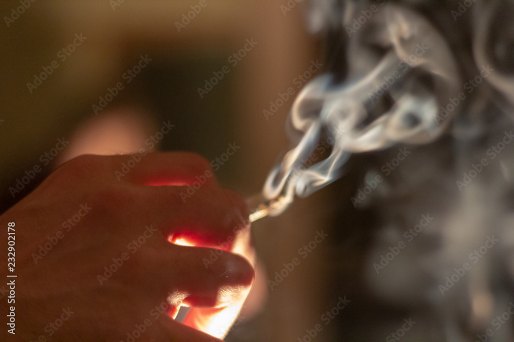 A hand holding a marijuana cigarette or joint with a large plume of thick smoke coming from it.