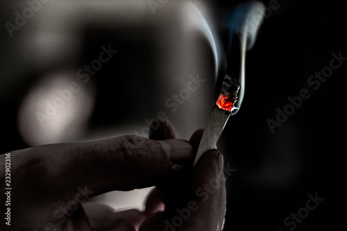 A dark lit scene of a hand passing a a joint with a bright red heater and a plume of smoke rising up.