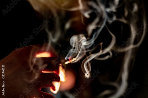 A hand holding a marijuana cigarette or joint in between two fingers as smoke swirls around.