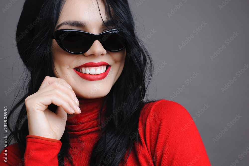 Naklejka premium Lovely young woman in stylish sunglasses smiling and looking away while standing on gray background. Portrait of beautiful young woman looking away.