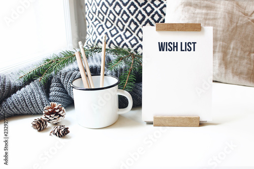 Winter desktop still life scene. Wish list card, board and wooden pencils in mug standing near window on white table background. Christmas concept. Pine cones and fir branch on wool plaid. photo