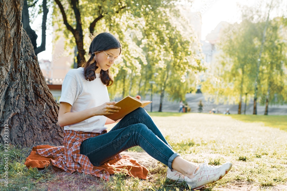 Girl student in glasses reading a book in the park sitting under a tree on the grass