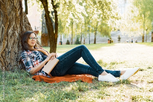 Beautiful woman with glasses reading book sitting on the grass near the tree in the park at sunset.