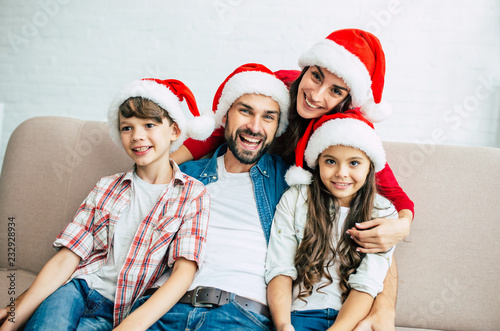 Merry Christmas  Happy young family in red santa hats are hugging each other and spending time in living room while sitting on the couch