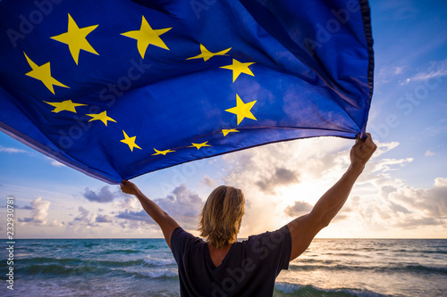 Man with blond hair holding EU European Union flag waving in the wind of a Mediterranean beach scene