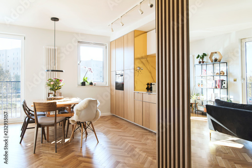 Stylish scandinavian open space with dining and living room with design sofa and family table. Bright and sunny room with big windows and brown wooden parquet. 