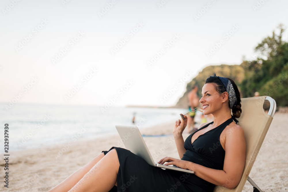 Portrait of a beautiful smiling young woman sitting on the beach with laptop.