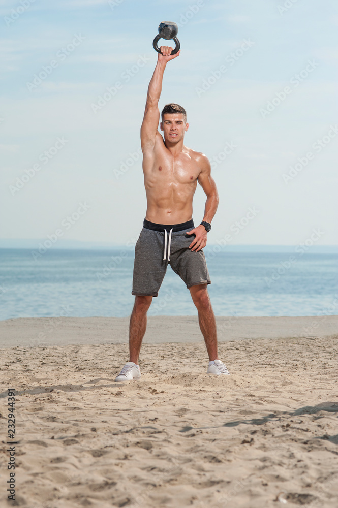 Athletic young handsome sweaty man short haircut with naked body holding  high up a kettlebell in one hand by the seaside in a daylight on a beach.  Stock Photo | Adobe Stock