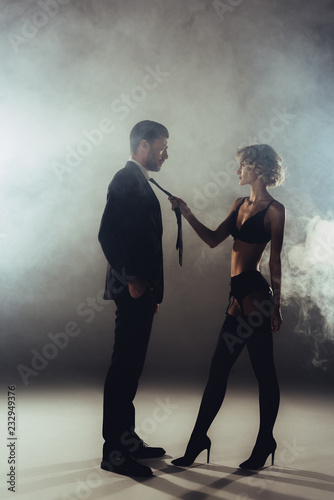 Fototapeta Blonde beautiful woman in lingerie pulling man by cravat on grey smoke backgroun