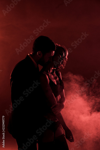 Man tenderly hugging beautiful woman and kissing her in neck on red smoke background